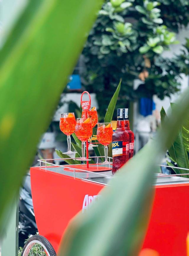 A red Aperol cart with glasses of spritz, framed by green leaves